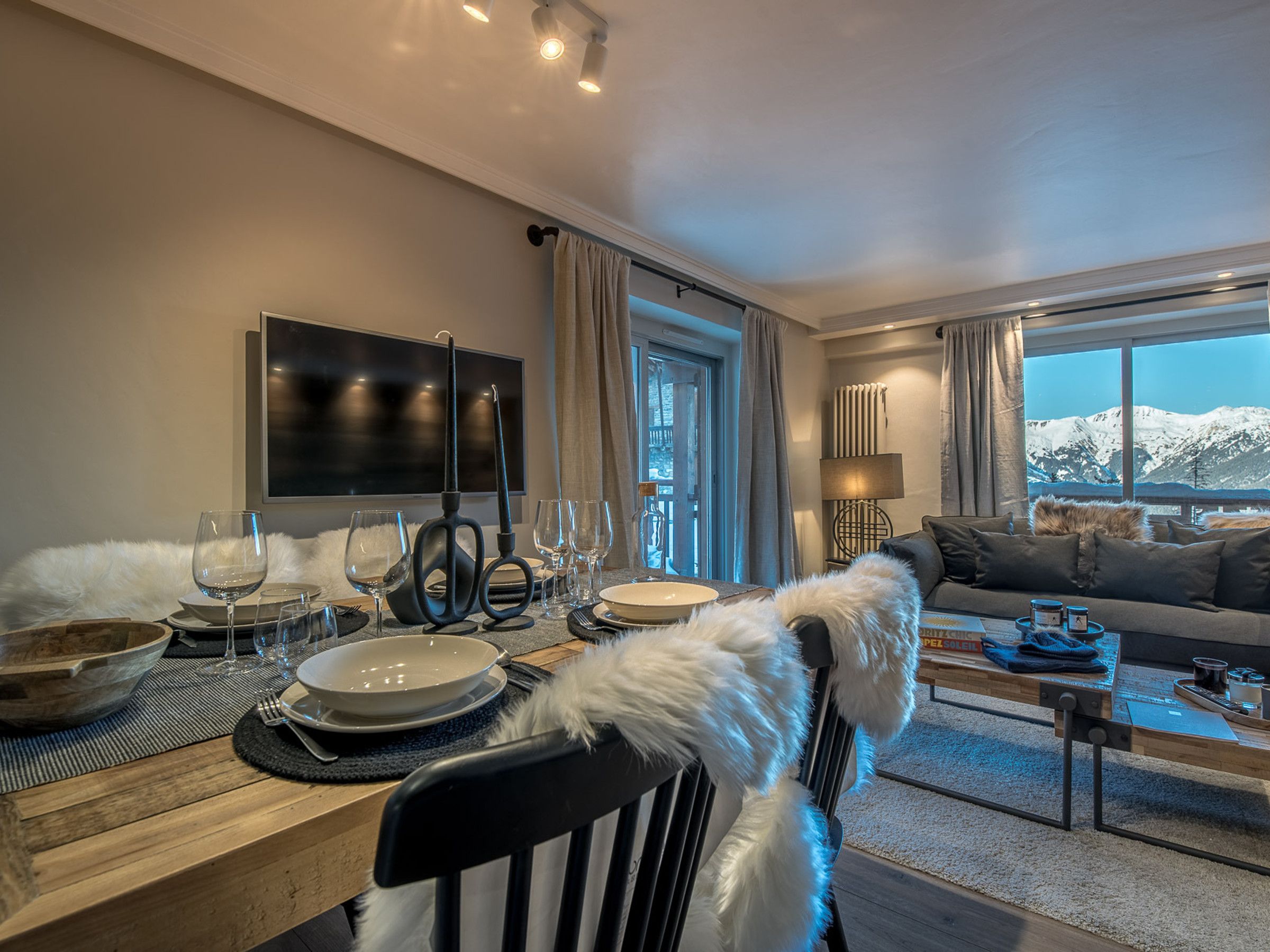 A dining area with a wooden table set for a meal, a sofa in the living room, and a view of snow-capped mountains through large windows.