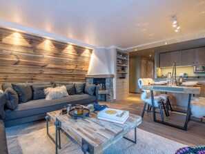 Ein modernes Wohnzimmer mit Sofa, Couchtisch und Kamin. Es gibt eine Küchenzeile mit Esstisch und Stühlen.