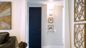 Una habitación con una puerta azul oscuro, una mesa de vidrio con una planta, un sofá gris y decoración en la pared.