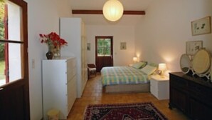 A bedroom with a bed, bedside table, lamp, dresser, and a window with a view of greenery. - Lovely rural house at Carcans close to lakes and surfing beaches (Carcans)