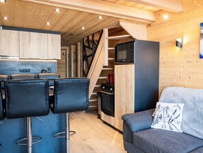 A modern kitchen with wooden cabinets, a built-in microwave, and a gas stove. There are two black bar stools and a gray sofa with a decorative pillow. - Renovated mountain apartment, 6 people, near slopes and amenities (Val Thorens)