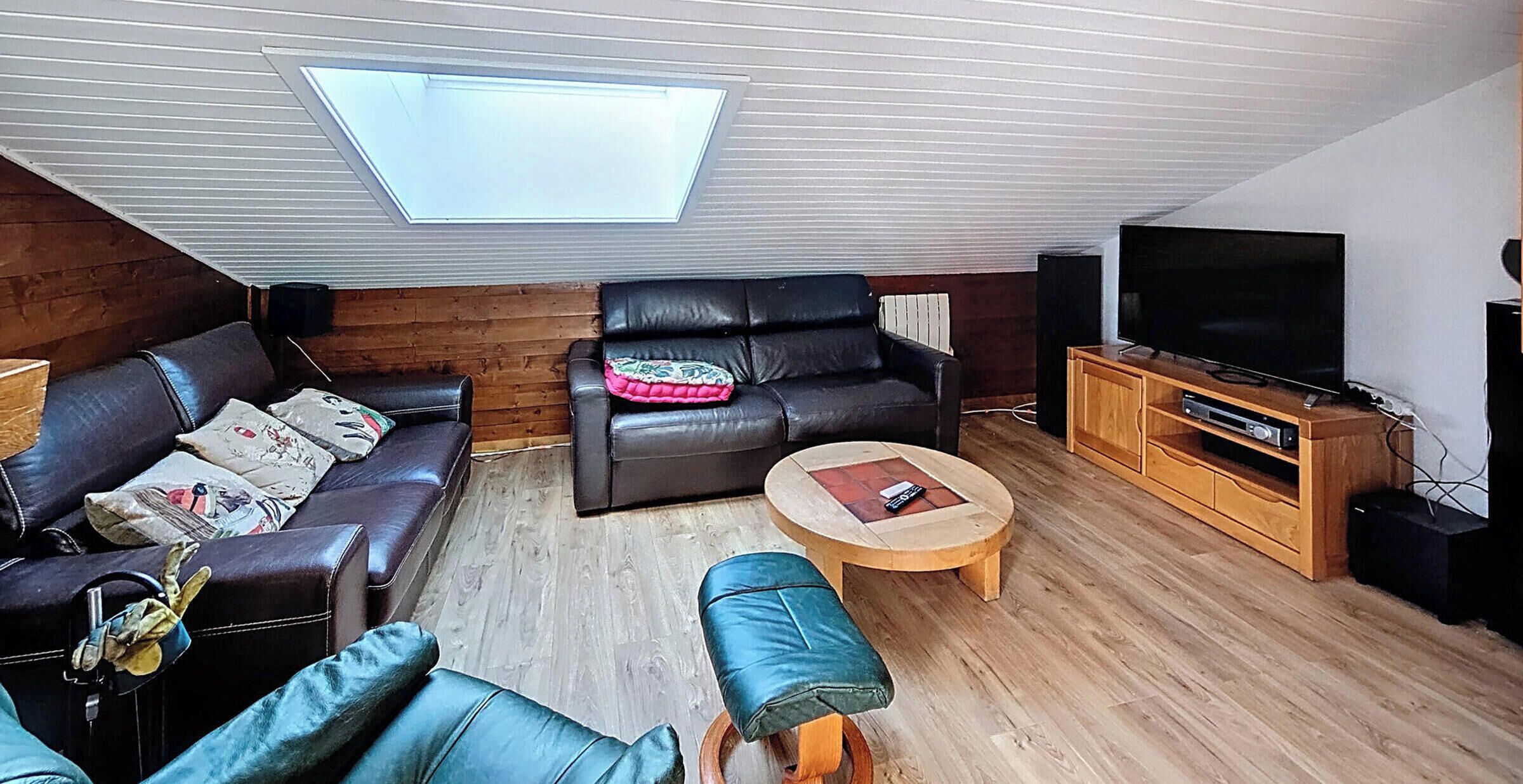 Una sala de estar acogedora con techo inclinado, un ventanal grande, un sofá de cuero, una mesa de centro de madera y un televisor sobre un mueble de madera.