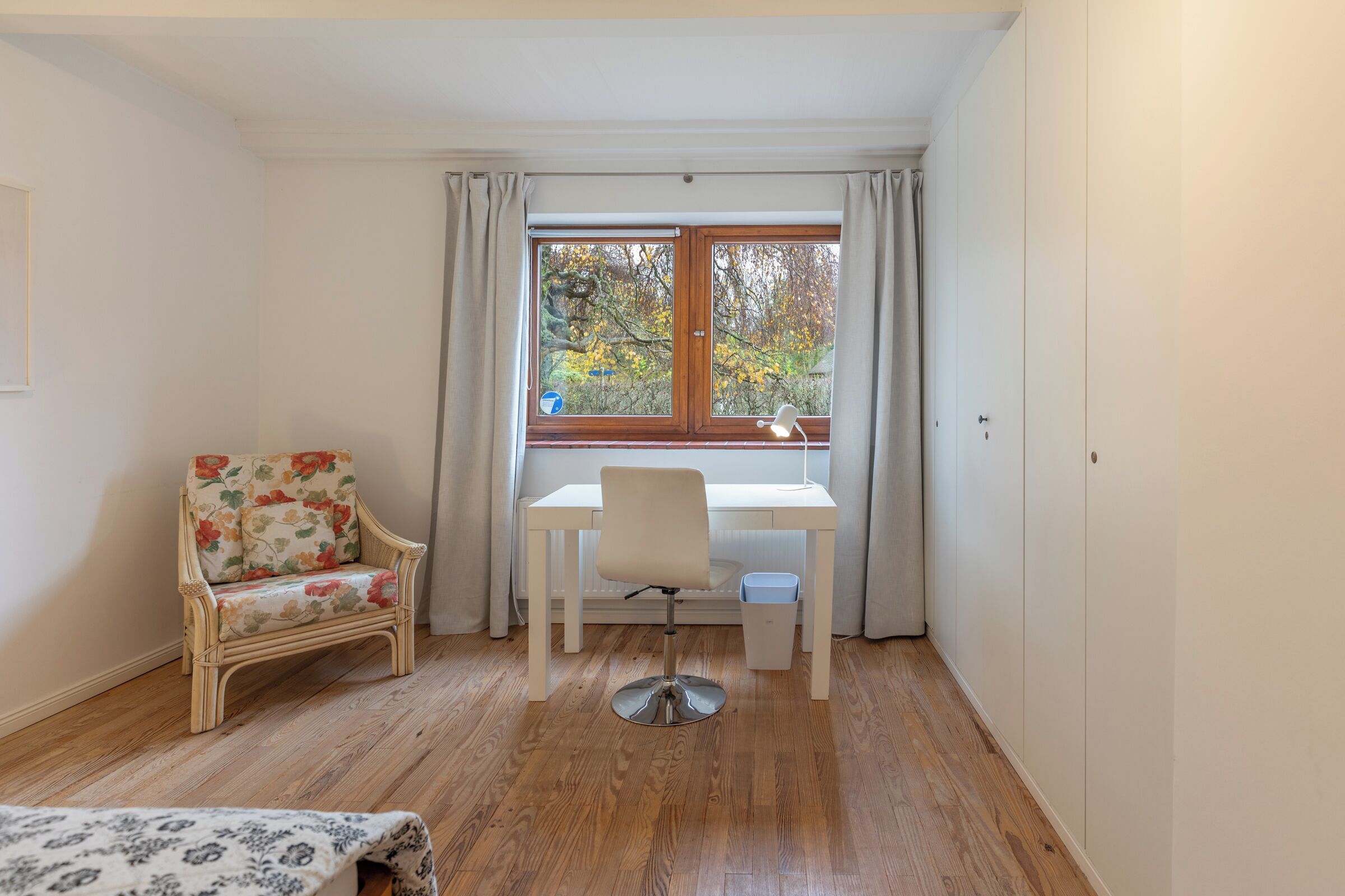 A room with a wooden floor, a white desk with a chair, a wicker chair, and a window with curtains.