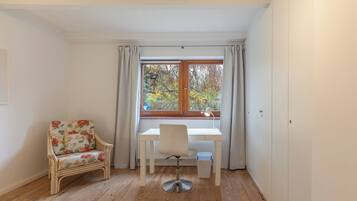 A room with a wooden floor, a white desk with a chair, a wicker chair, and a window with curtains.