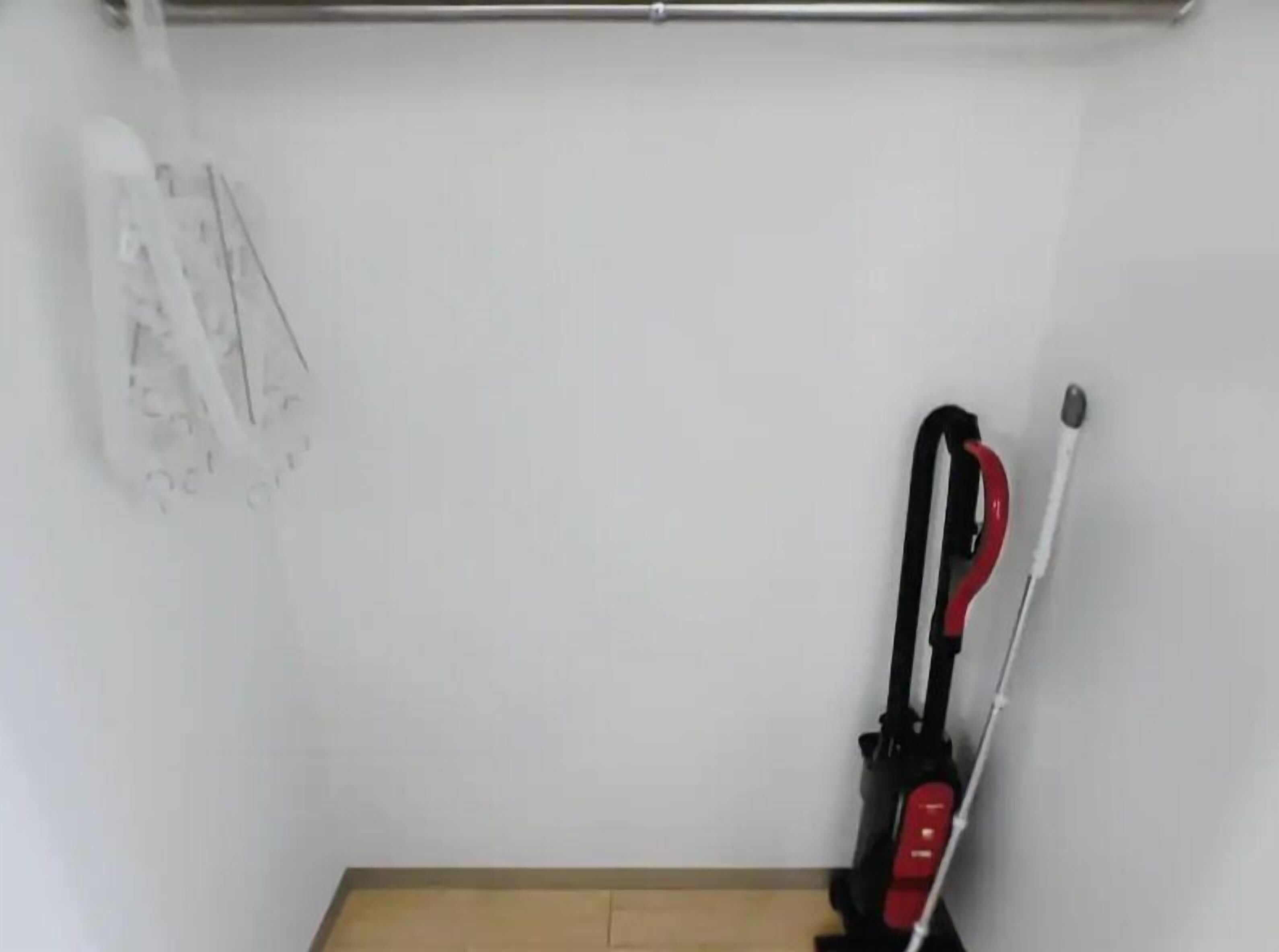 A room with a mop and broom leaning against the wall, a wooden floor, and a white wall.