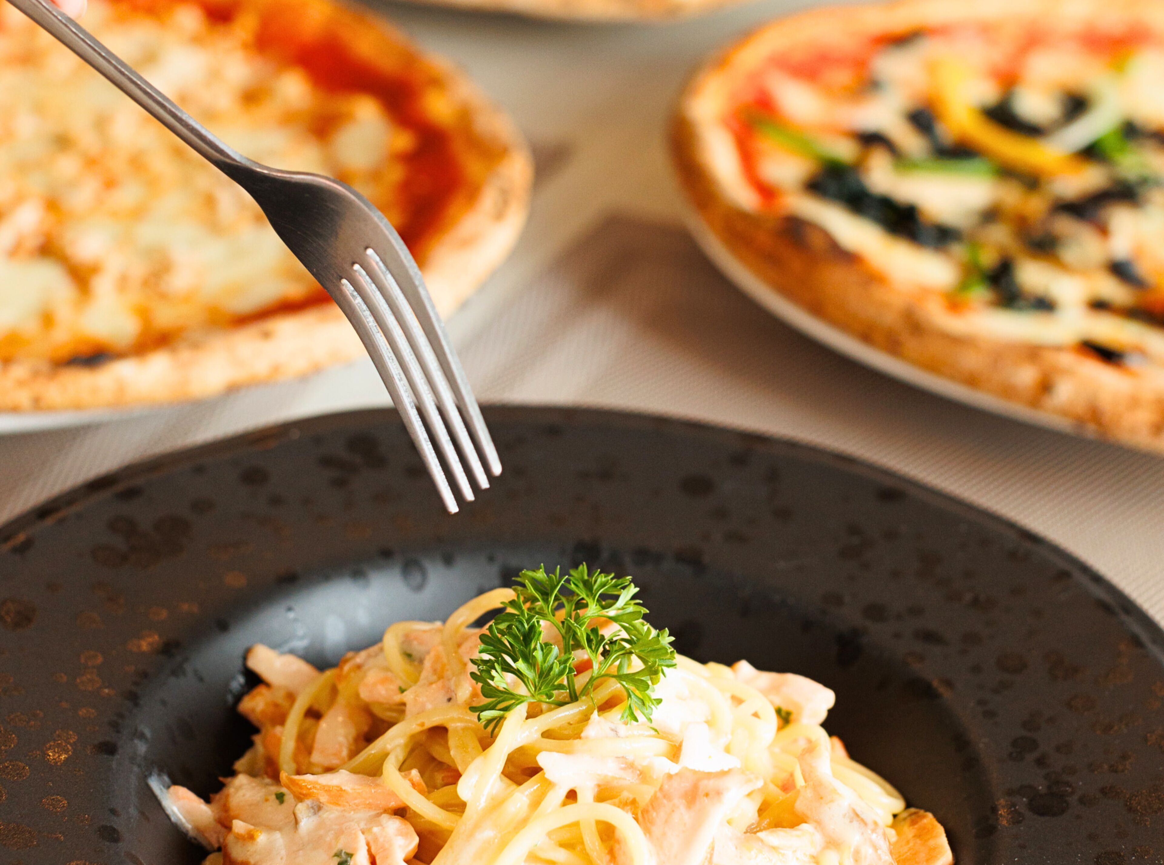 Een bord pasta, versierd met peterselie, een vork en twee pizza's op de achtergrond.