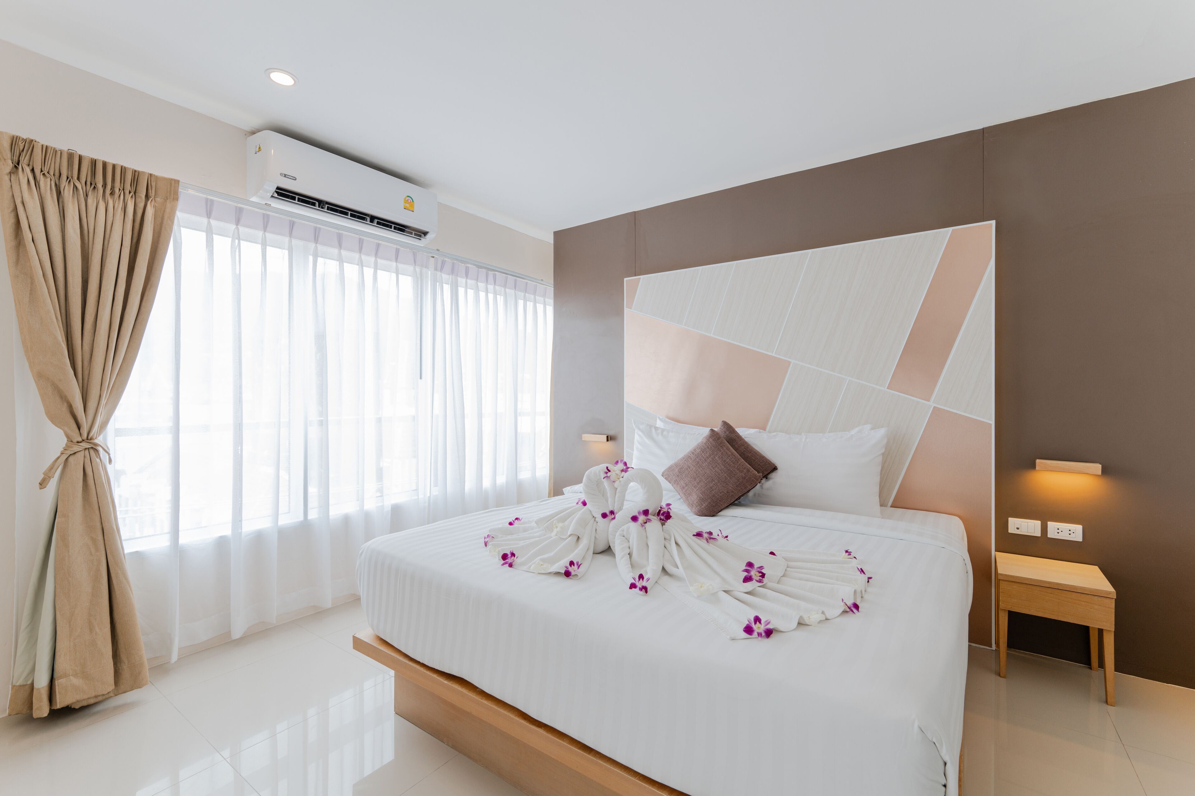 A hotel room with a large bed, a headboard with geometric patterns, a bedside table, and a window with sheer curtains.