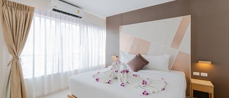 A hotel room with a large bed, a headboard with geometric patterns, a bedside table, and a window with sheer curtains.