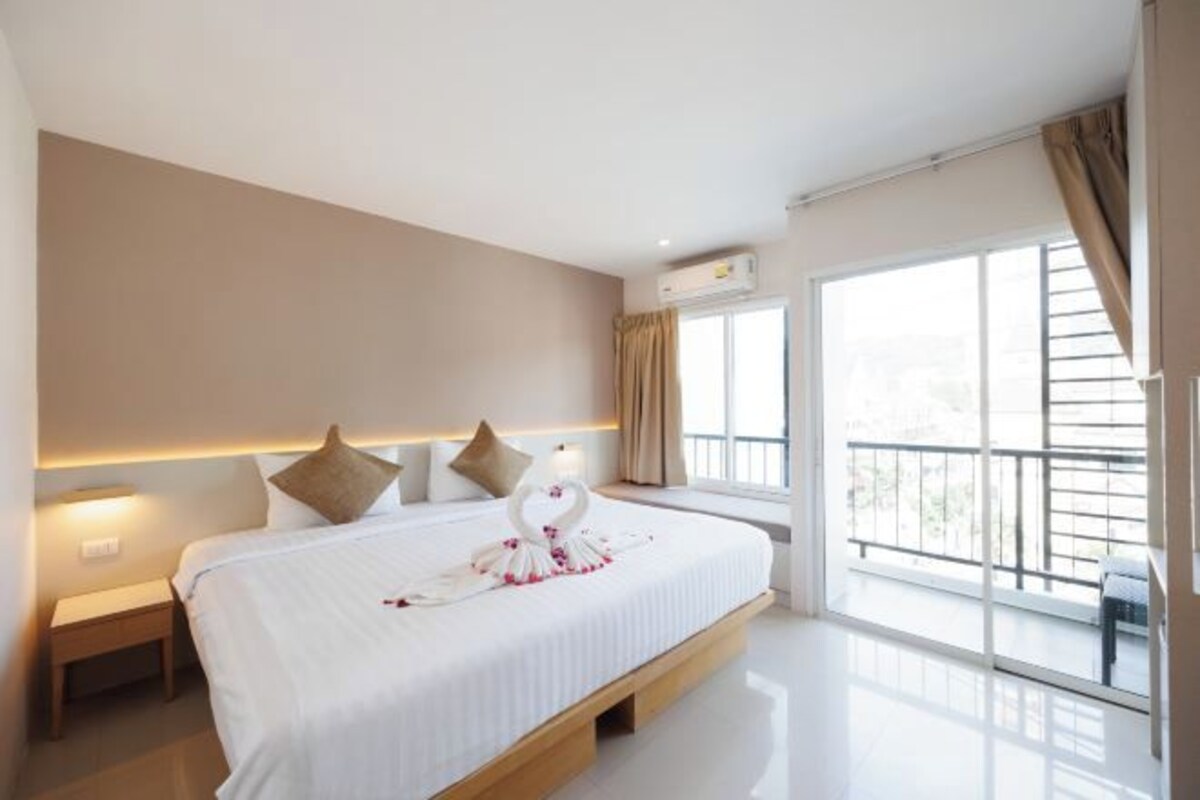 Quarto de hotel com cama grande, mesa de cabeceira, janela com cortinas e varanda com guarda-corpo.