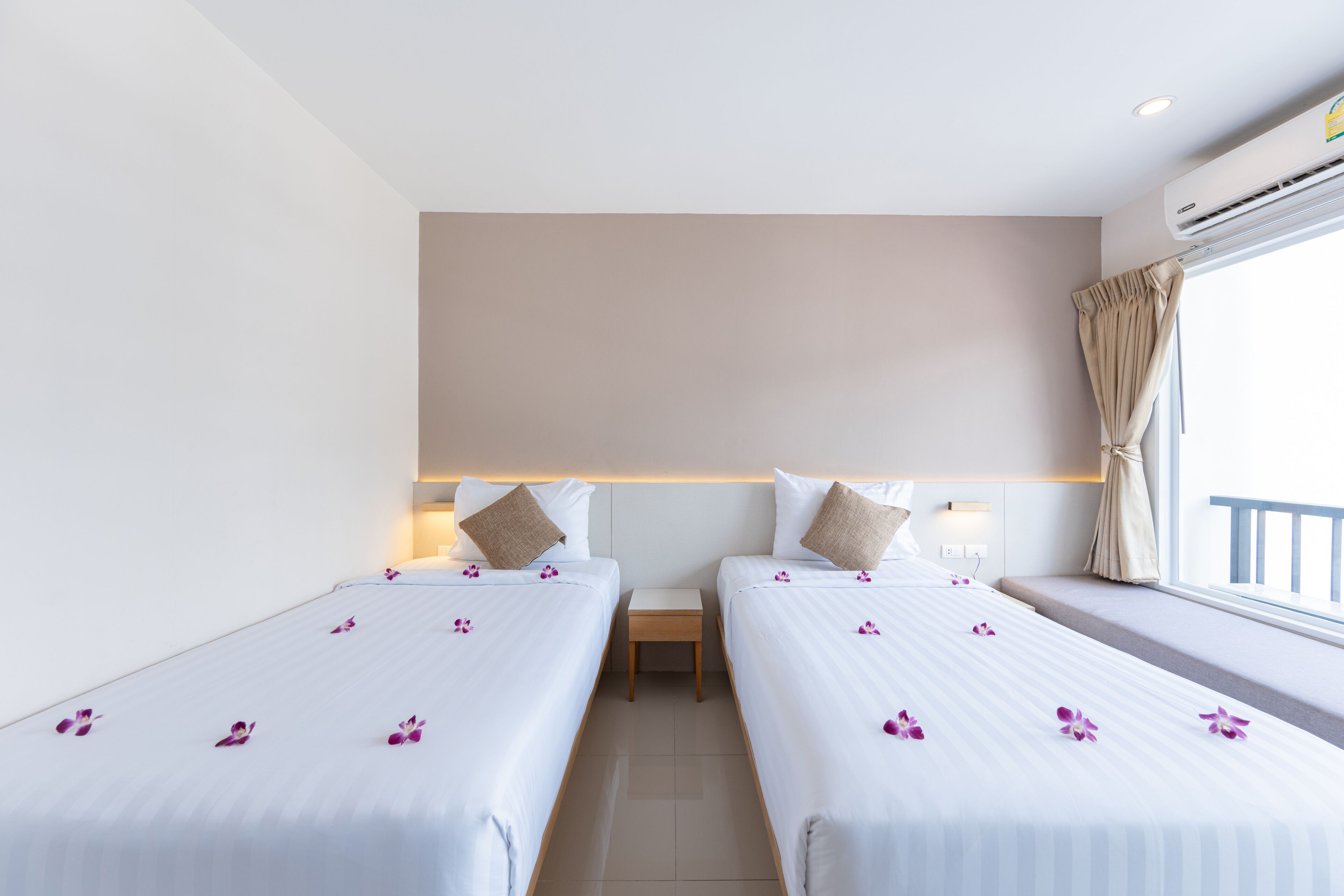 Habitación de hotel con dos camas individuales, una mesita de noche pequeña, un equipo de aire acondicionado y una ventana con cortinas.