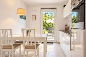 Eine moderne Küche mit weißen Schränken, ein Essbereich mit weißem Tisch und Stühlen und ein Blick ins Freie durch eine Glastür.