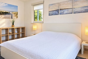 Ein Schlafzimmer mit einem großen Bett, einem Holfnachtkästchen, einem Nachttisch, einem Fenster mit Jalousien und Wandbildern.