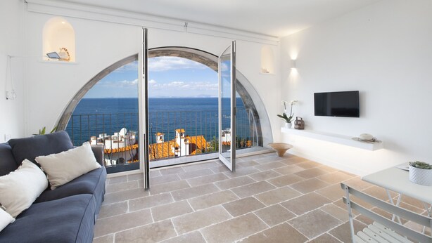 A modern living room with a large arched window offering a view of the sea and buildings.