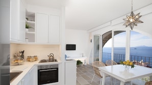 A modern kitchen with white cabinetry, a built-in oven, and a dining area with a white table and wicker chairs. A star-shaped light fixture hangs above the table, and a large window offers a view of the sea.