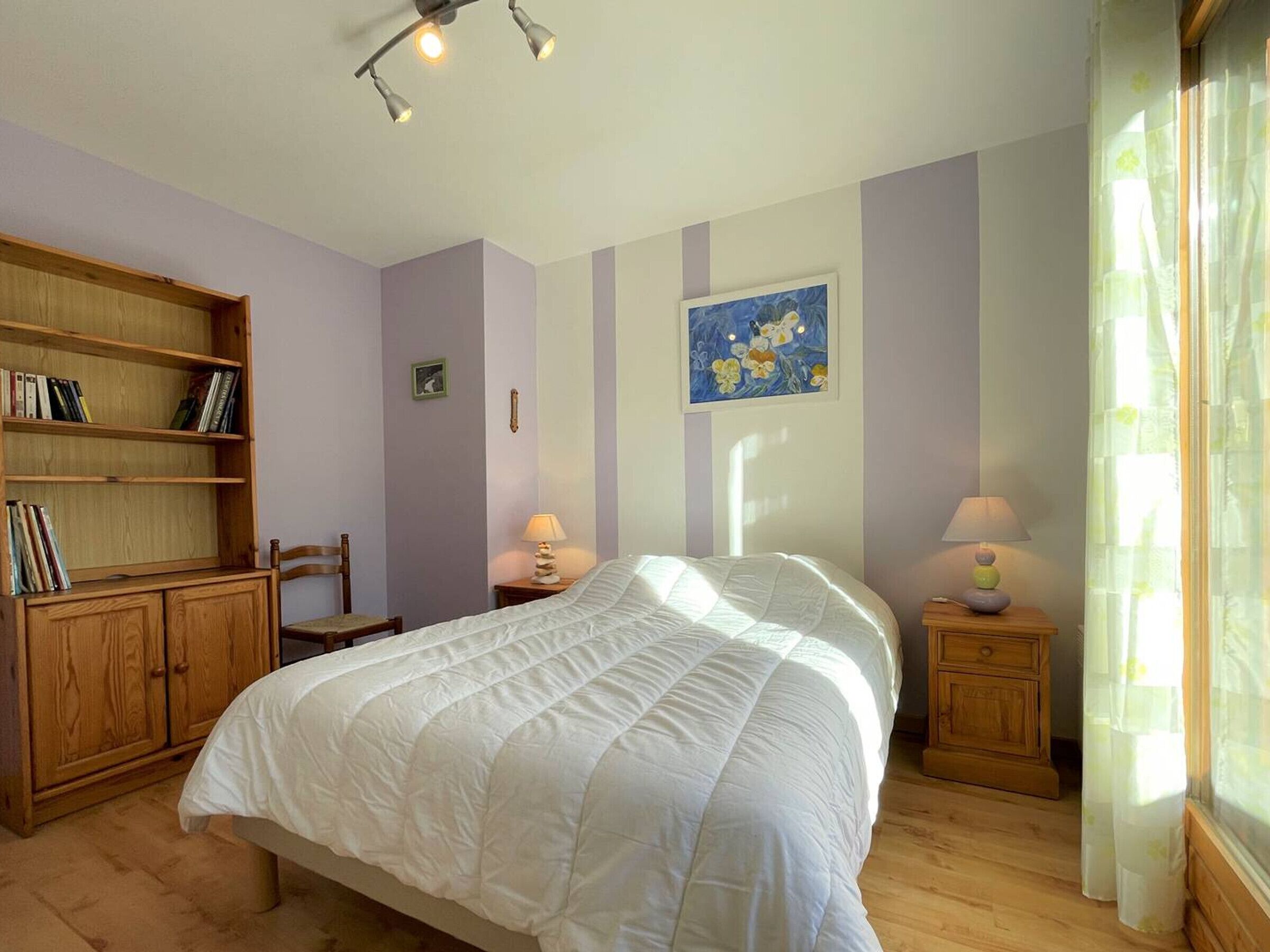 A bedroom with a bed, wooden bookshelf, two bedside tables with lamps, a chair, and a window with curtains.