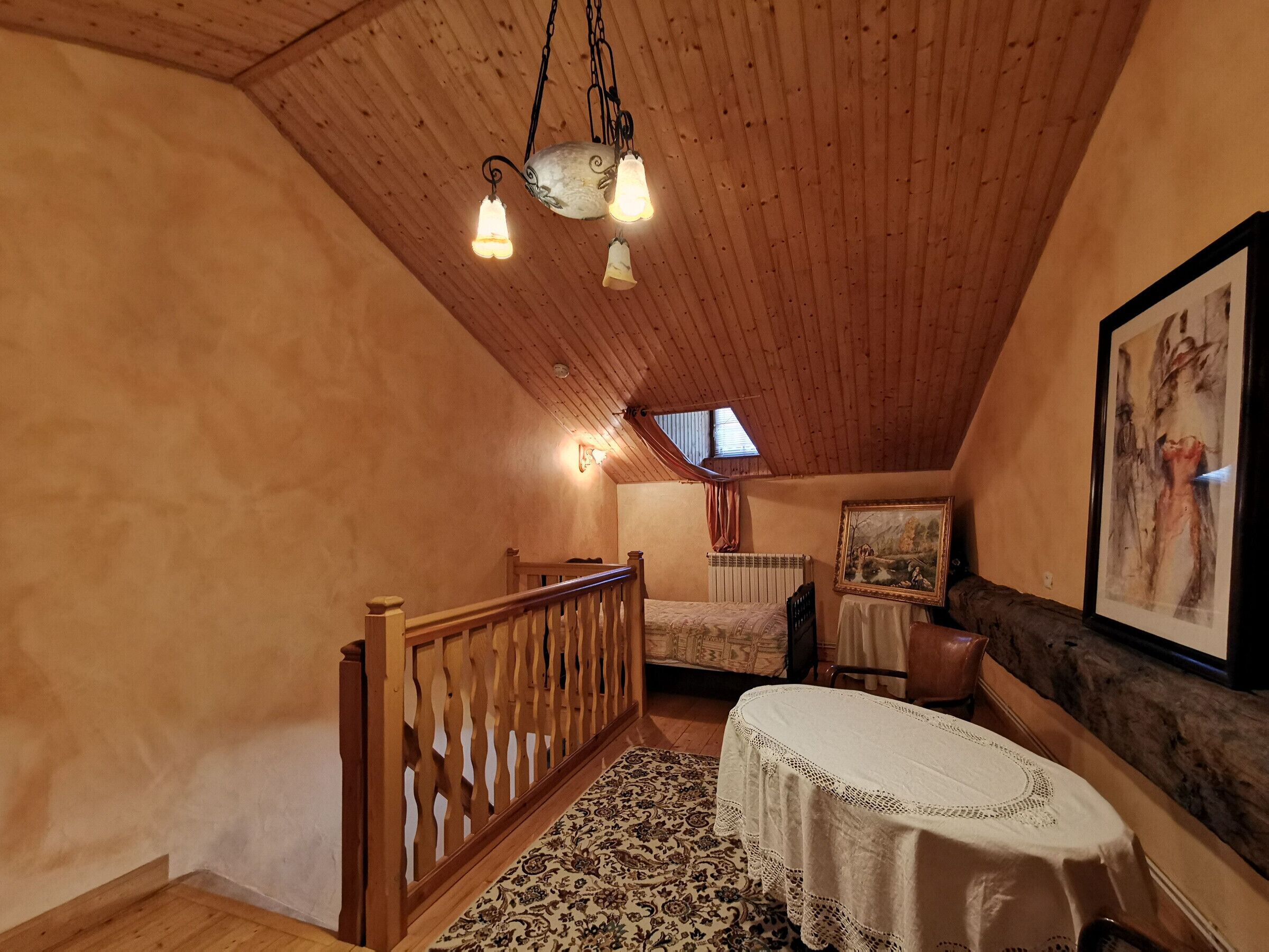 Ein gemütliches Schlafzimmer im Dachgeschoss mit Holzbalkendecke, einem Bett mit gemustertem Überwurf, einem kleinen Tisch mit Spitze und einem Bild an der Wand.
