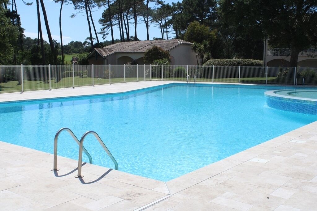 A rectangular swimming pool with a tiled deck, metal handrails, and a white fence surrounding it.