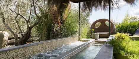 A tranquil outdoor pool area with a stone-lined water feature, surrounded by lush greenery and a wooden pergola.