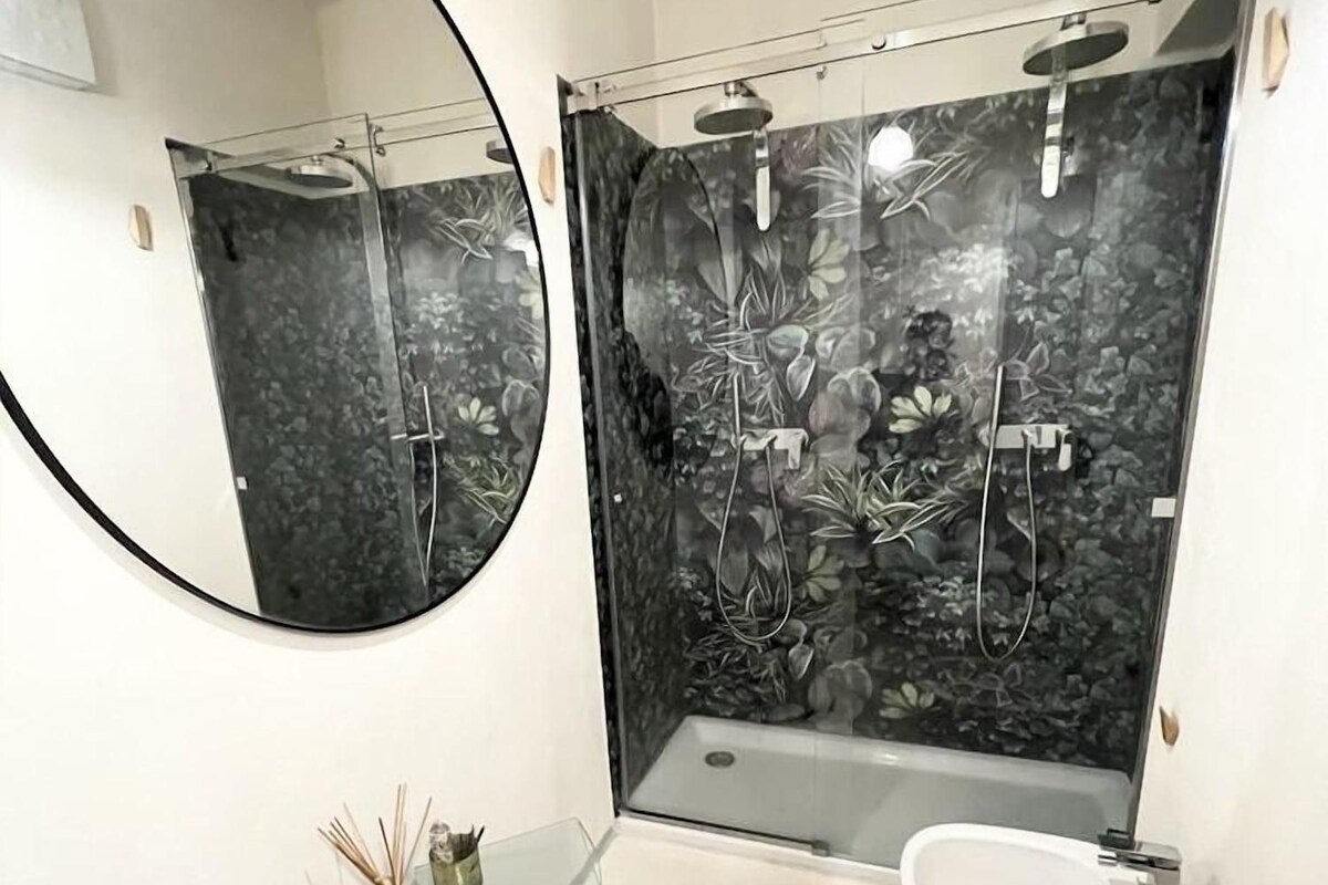 A modern bathroom with a round mirror, a glass side table, a bathtub, and a toilet.