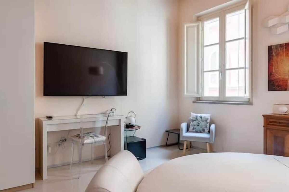 A modern bedroom with a large bed, a TV mounted on the wall, a desk with a chair, and a window providing natural light.