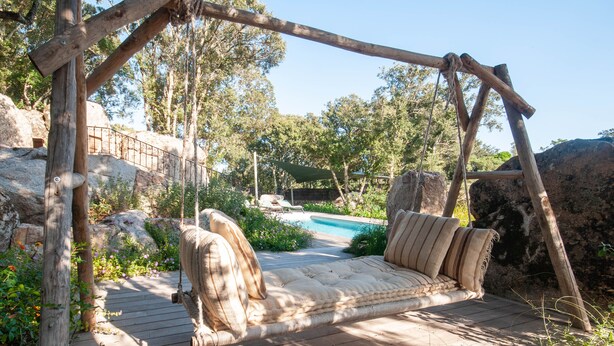 Un fauteuil suspendu en bois avec des coussins, une terrasse en bois, une piscine et de la verdure environnante.