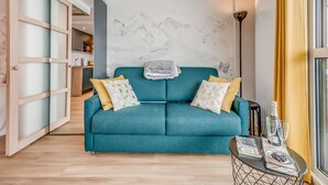 Ein blaugrünes Sofa mit gemusterten Kissen, ein kleiner Tisch mit einer Flasche und Gläsern sowie eine Wand mit Bergmotiv.
