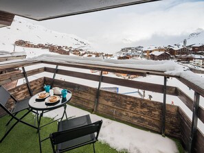 A balcony with a table and chairs, overlooking a snowy mountain village with buildings and a ski lift.