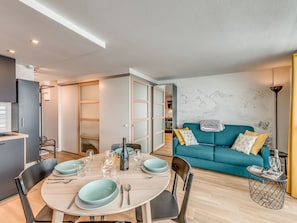 A modern living room with a teal sofa, a dining table set for four, and a kitchen area visible through an open door.