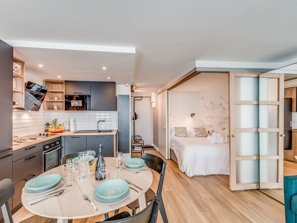 A modern kitchen with a dining area set for four, a bed with a view, and a sliding door to a bedroom.