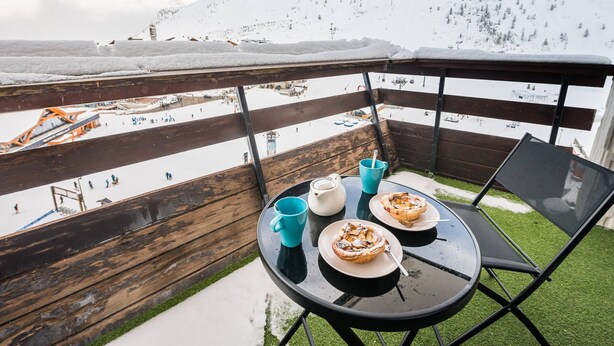 Ein Balkon mit Tisch für zwei Personen, mit Blick auf einen verschneiten Berg und eine Skipiste.