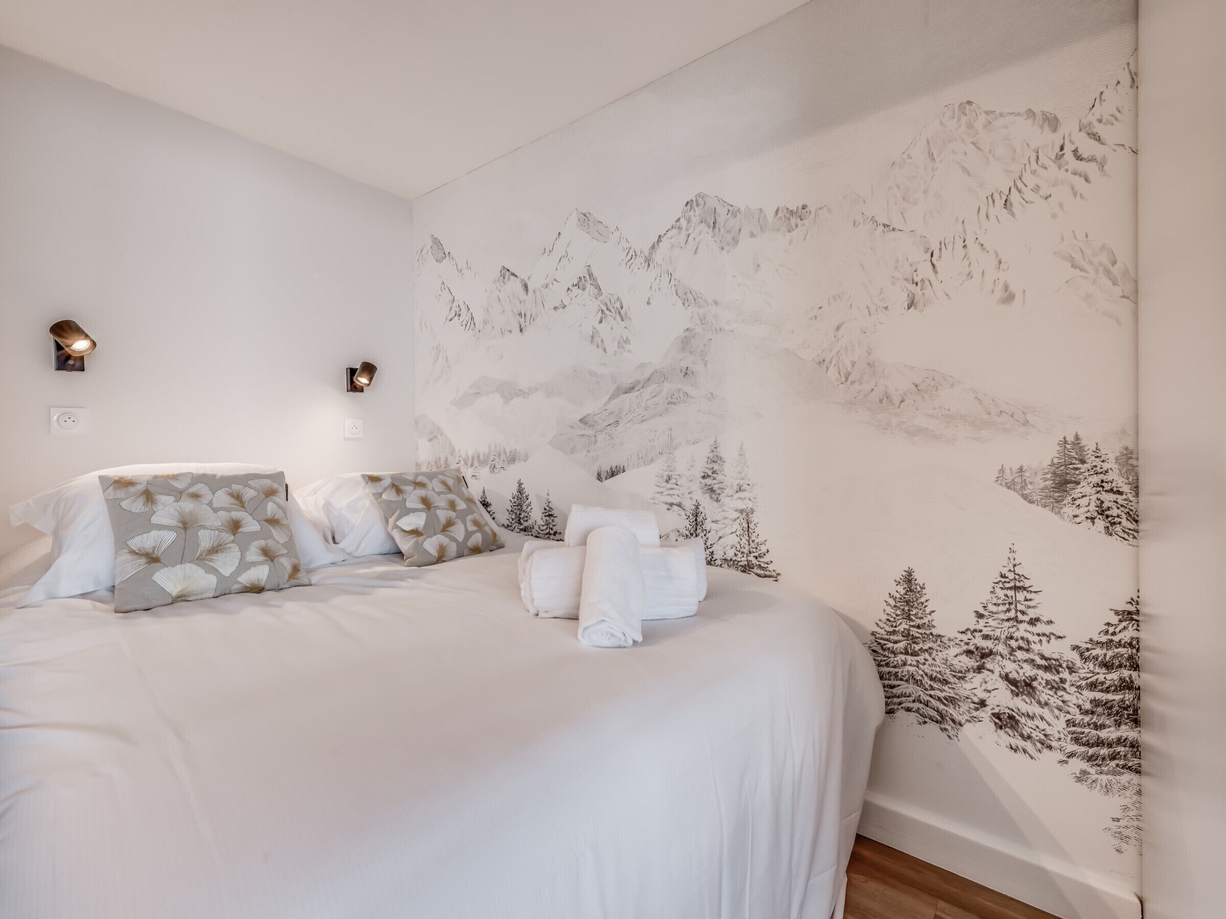 Un dormitorio con papel tapiz alusivo a las montañas, una cama bien hecha con ropa blanca y dos lámparas fijadas a la pared.