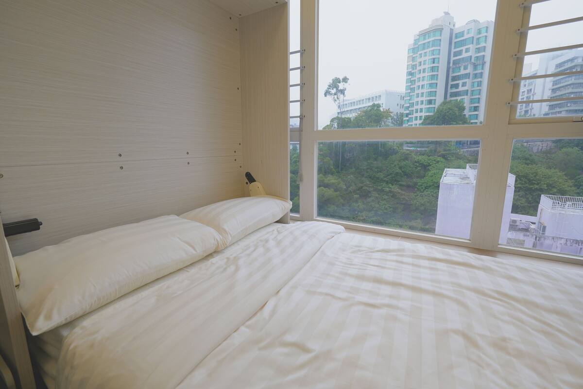 A bedroom with a bed, a window, and a view of buildings and greenery.
