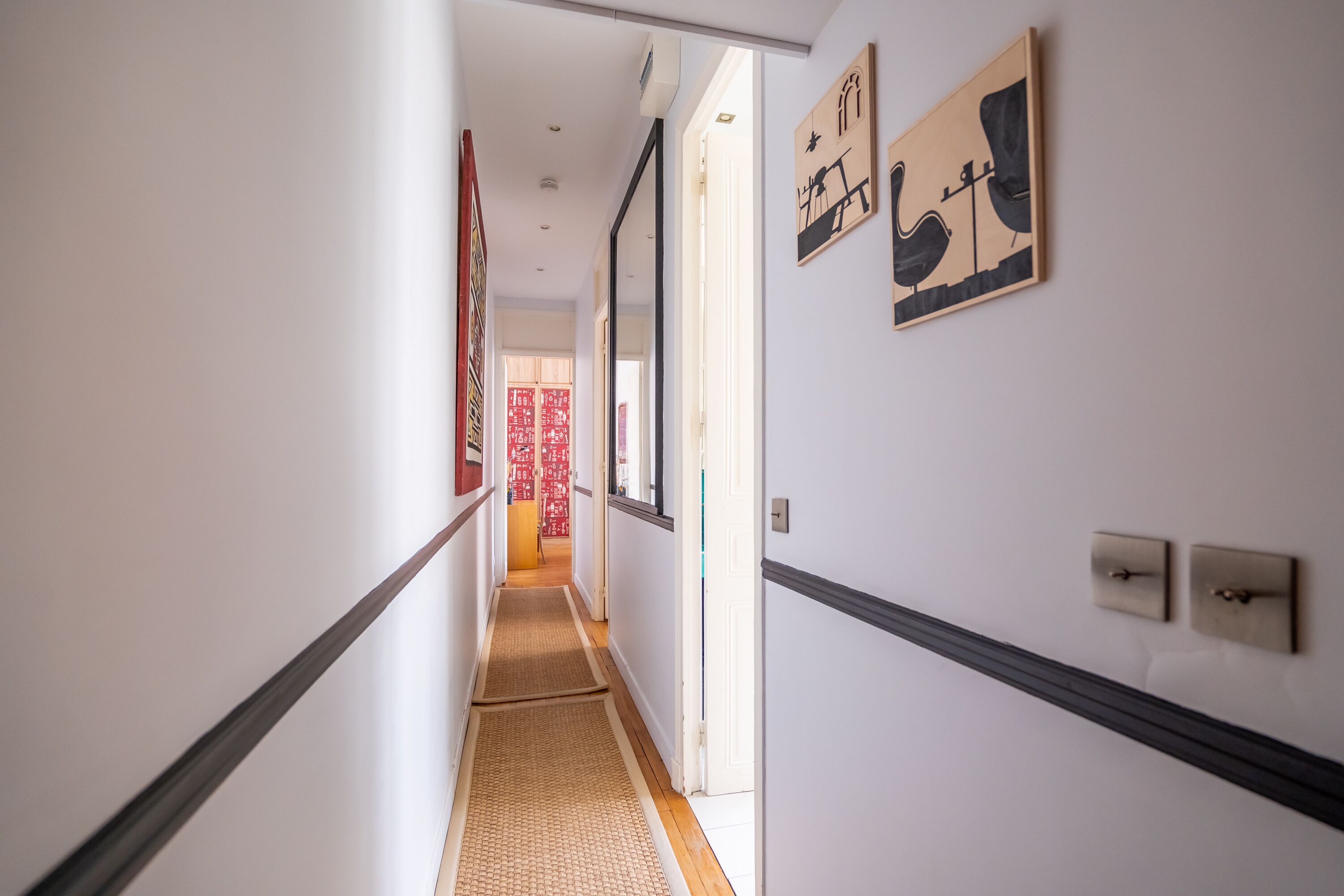 A hallway with a wooden floor, white walls, and black trim. There are framed artworks on the walls and a door at the end.