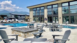 A pool area with lounge chairs and tables, a fire pit, and a modern building with large windows.