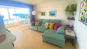 A living room with a teal sofa, colorful cushions, a small table, and a chair. There are two framed pictures on the wall, a hanging light fixture, and a plant on a shelf. - Salinetas, first line of the sea and beach for 4 people (telde)