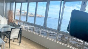 A room with a large window overlooking a coastal area, a glass table with chairs, and a view of the sea and buildings. - Salinetas, first line of the sea and beach for 4 people (telde)