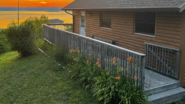 Una casa de madera con un porche, vista al atardecer sobre un cuerpo de agua y un área verde con flores.