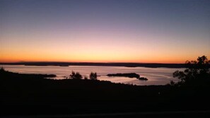A sunset view over a calm body of water with distant landforms and trees silhouetted against the sky.