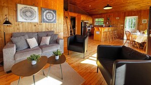 Interior de una cabaña de madera con sofá, sillones, zona de comedor y cocina.