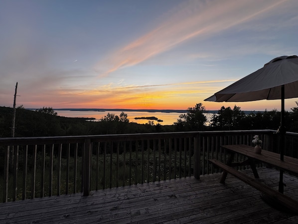 A wooden deck with a picnic table and umbrella, overlooking a body of water at sunset.