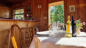 Interior de una cabaña de madera con una mesa de comedor para dos, una barra y un ventanal con vistas a árboles.