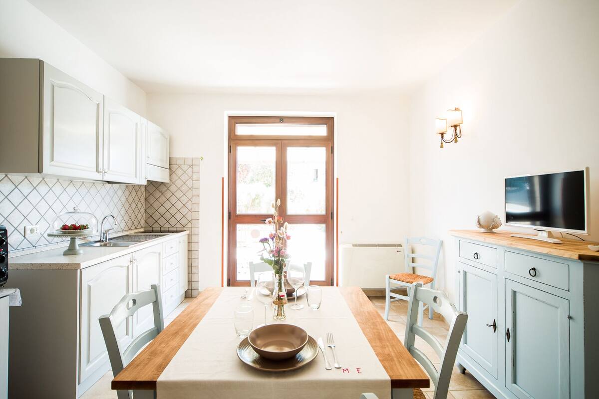 Una cocina luminosa con armarios blancos, una mesa de comedor de madera dispuesta para dos personas y un televisor a la derecha.