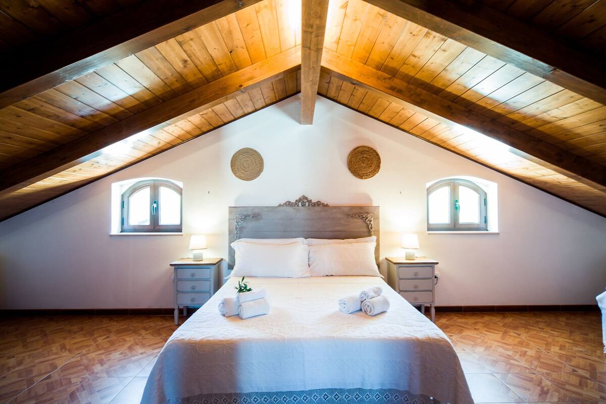 Un dormitorio con techo inclinado, dos mesitas de noche, una cama con ropa de cama blanca y dos ventanas con arcos.