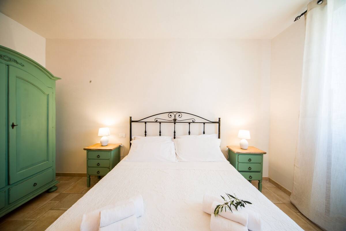 Una cama bien hecha con sábanas blancas, dos mesitas de noche con lámparas y un armario verde.