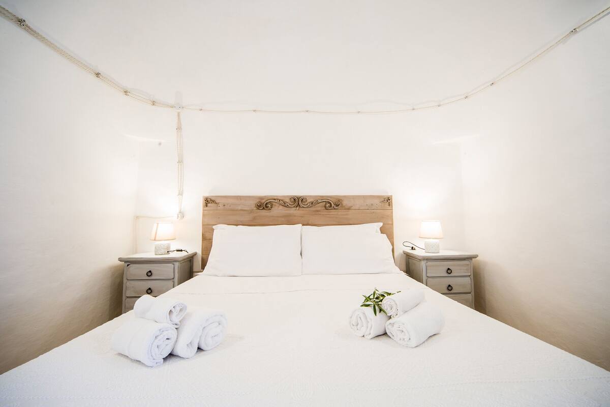 Una cama bien tendida con sábanas blancas, dos mesitas de noche con lámparas y un cabecero de madera.