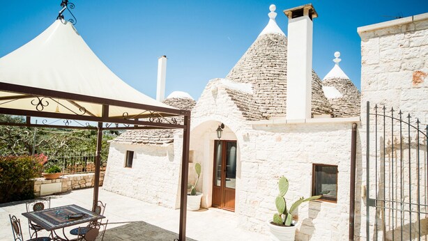 Un edificio de piedra blanca con techo cónico, un patio cubierto con una mesa y sillas, y una planta de cactus.
