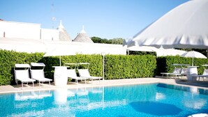 Una piscina con sillas de descanso y sombrillas, rodeada por un seto y un edificio con fachada blanca y una torre.