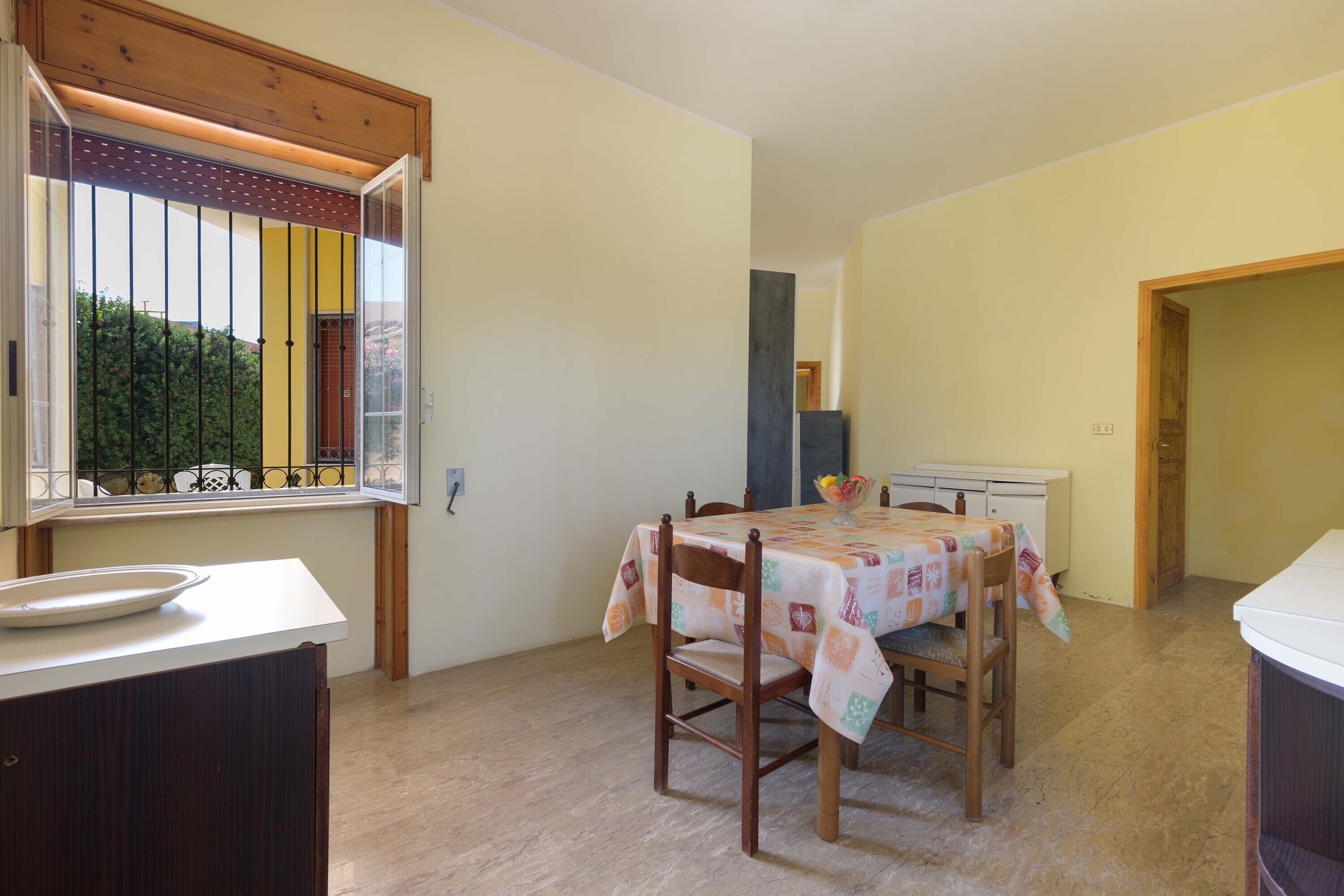 Un comedor con una mesa lista para dos personas, una ventana con vistas a follaje, y una zona de cocina con armarios y una encimera.