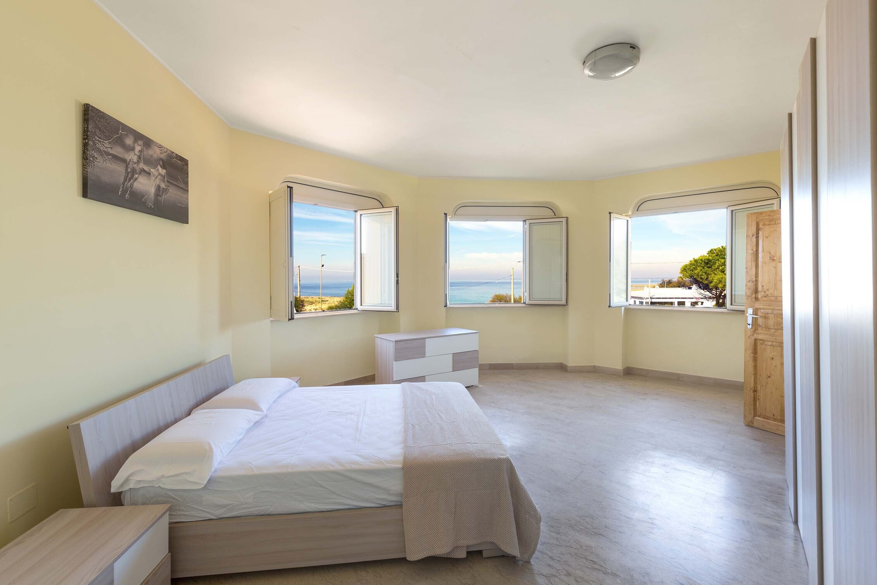 Ein Schlafzimmer mit Bett, Nachttischen, einem Schminktisch und großen Fenstern mit Blick auf das Meer.