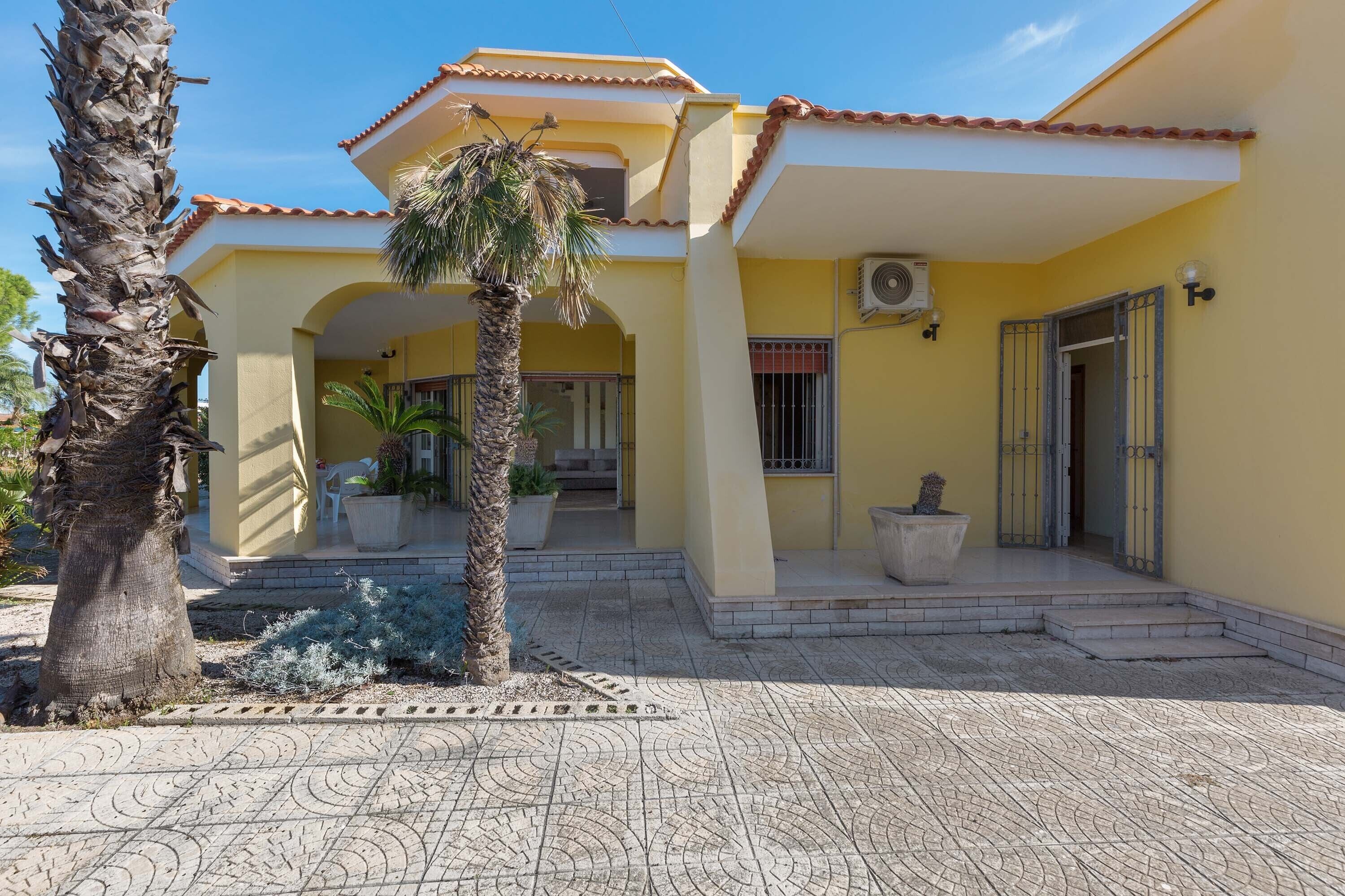 A yellow building with a tiled patio, palm trees, and potted plants.